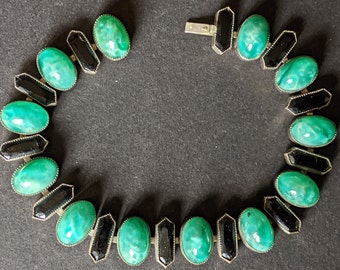 Puede incluir: Un collar verde y negro con piedras verdes ovaladas y detalles negros. El collar está hecho de metal y tiene un cierre.