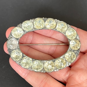 May include: An oval-shaped silver brooch with a row of clear faceted glass stones set in a silver frame.