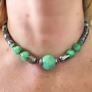 May include: A green and silver necklace with a large green bead in the center. The necklace is made of small silver beads and green beads with a marbled pattern.