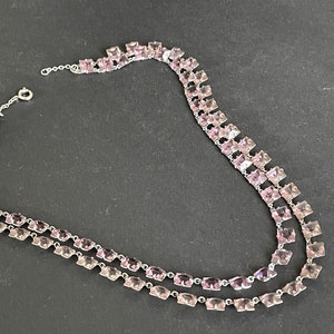 Puede incluir: Un collar de doble cadena con cadena de plata y piedras cuadradas de color rosa.