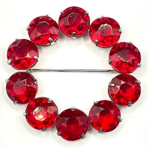 May include: A red rhinestone brooch in the shape of a circle. The brooch is made of 10 round red rhinestones set in silver.