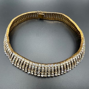 May include: A gold-coloured choker necklace with a row of sparkling clear rhinestones. The necklace has a flexible design with a clasp closure. The jewellery is displayed on a dark surface.