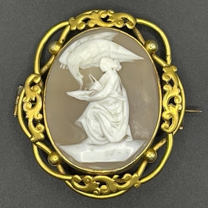 Può includere: Una spilla a cammeo ovale con una figura bianca scolpita di una persona in ginocchio che tiene un recipiente sotto un grande uccello con le ali spiegate, su uno sfondo marrone chiaro all'interno di una cornice in metallo dorato lucido.