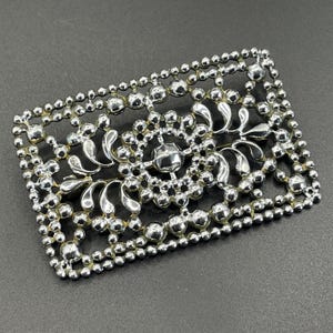 May include: A rectangular silver brooch with an elaborate floral pattern. The brooch has a central crystal-like detail and is framed by a border of small, round beads. The intricate design and polished finish give it a vintage appearance.