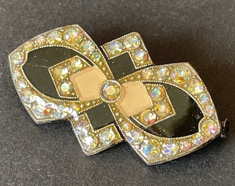 Könnte beinhalten: Eine geometrische Brosche in Schwarz und Weiß mit schillernden Strasssteinen. Die Brosche hat ein zentrales Design aus rosa Emaille.