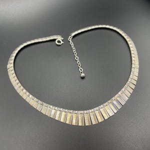 Könnte beinhalten: Eine silberfarbene Choker-Halskette mit einem strukturierten, geometrischen Design. Die Halskette besteht aus einer Reihe rechteckiger Segmente, die zu einem flexiblen Band verbunden sind. Sie hat einen Hakenverschluss und eine verstellbare Kette. Der Hintergrund ist schwarz.