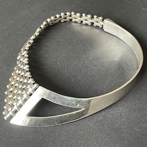 Könnte beinhalten: Eine silberne Choker-Kette mit geometrischem Design. Die Kette besteht aus einer gewebten Metallkette und hat ein glattes, gebogenes Band.