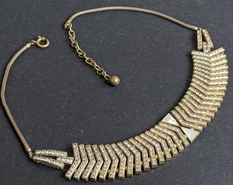 May include: A gold tone necklace with a geometric design featuring a row of clear rhinestones. The necklace has a chain with a small ball at the end.