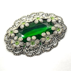 Peut inclure: Broche ovale avec une grande pierre vert émeraude facettée au centre. La pierre est entourée d'un cadre filigrané argenté et ornée de petites fleurs argentées avec des centres verts. La broche a une esthétique vintage.