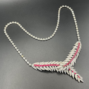 Op de afbeelding: Een zilverkleurige ketting met een bladachtig ontwerp. De ketting heeft heldere, ronde edelstenen en een rij rode edelstenen. De ketting wordt gepresenteerd op een donkere ondergrond.