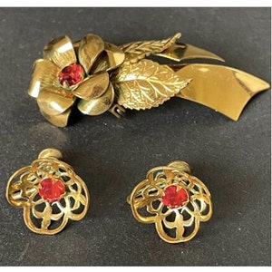 Könnte beinhalten: Eine goldfarbene Blumenbrosche mit roten Strasssteinen und passenden Ohrringen. Die Brosche zeigt eine große Blume mit Blättern und einem Band. Die Ohrringe sind klein und zart.