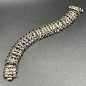 May include: A silver chain bracelet with a detailed, intricate design. The bracelet is made of linked, interlocking pieces that create a textured, patterned look.