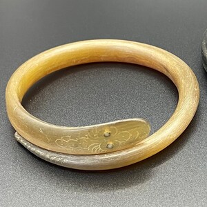 May include: A translucent, light tan, coiled bangle bracelet shaped like a snake. The surface features a smooth, polished texture with subtle grain patterns. The head of the snake is engraved with a floral design and two small, white circular inlays.