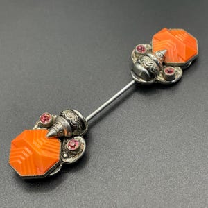 Peut inclure: Épingle à cravate vintage en argent avec une pierre géométrique orange et de petites pierres roses. L'épingle a un design argenté décoratif et est posée sur un fond sombre. Un bijou unique.