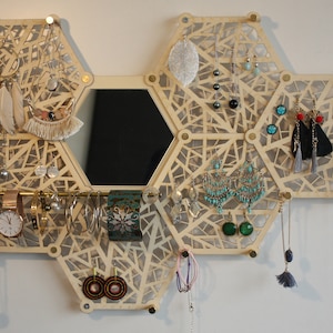 Peut inclure: Un organisateur à bijoux hexagonal en bois avec un miroir au centre. L'organisateur comporte plusieurs compartiments pour les boucles d'oreilles, les colliers et les bracelets. L'organisateur est en bois clair avec une finition naturelle.