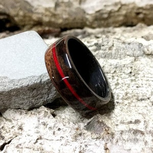 May include: A black wooden ring with a red stripe inlay. The ring is sitting on a piece of bark.