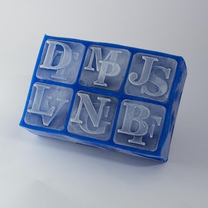 May include: A blue silicone ice cube tray with ice cubes in the shape of letters. The letters are D, M, J, S, L, N, and B. The tray is rectangular and has six compartments.