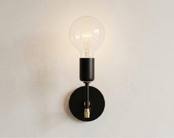 May include: A wall sconce featuring a matte black circular backplate, a slender black arm, and a cylindrical black socket. A large, clear, round glass light bulb is installed in the socket, emitting a warm glow from its visible filament.