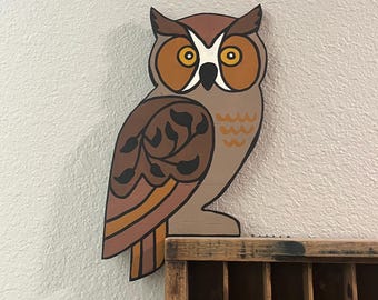 Pode incluir: Um recorte de madeira em forma de coruja em tons de castanho, bege e laranja, com padrões de folhas pretas na asa. Abaixo da coruja encontra-se uma caixa de tipografia de madeira com compartimentos, contendo uma pequena caixa amarela com o rótulo LOVE STRIKES.