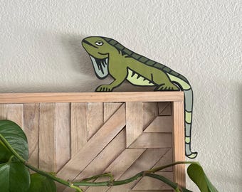 Pode incluir: Um recorte plano de iguana verde com contornos pretos e uma cauda longa e curva que pende sobre a borda de uma moldura de madeira. A iguana apresenta uma barriga verde-clara, costas verde-escuras e uma papada distinta sob o queixo.