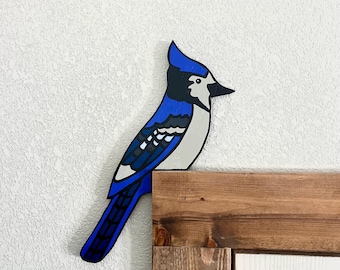 Pode incluir: Uma decoração de madeira recortada em forma de gaio-azul, com detalhes pintados em tons vibrantes de azul, preto, branco e cinzento. A ave é representada de perfil com uma crista na cabeça, pousada no canto de uma moldura de porta.
