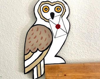 Pode incluir: Uma decoração de parede plana em forma de coruja branca com grandes olhos amarelos, bico preto e asas com manchas castanhas. A coruja segura no bico um envelope branco, selado com um selo de cera vermelho com um símbolo.