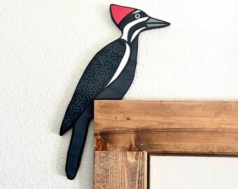 Pode incluir: Um recorte decorativo plano de madeira de um pica-pau com corpo preto, um padrão geométrico na asa, uma faixa branca no pescoço e uma crista vermelha proeminente na cabeça. A ave foi concebida para pousar sobre uma moldura de porta.