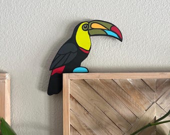 Pode incluir: Uma decoração de parede em forma de tucano com corpo preto, peito amarelo brilhante e um bico grande e multicolorido com seções em verde-oliva, vermelho e azul-claro. A ave tem um pequeno detalhe azul-claro e uma pena vermelha na cauda.