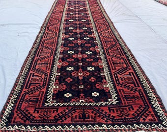 May include: A long, rectangular rug with a detailed geometric pattern. The rug features a dark red and black central field with repeating floral and geometric motifs. The borders are red and black with white accents and fringe.