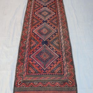 May include: A long, narrow rug with a geometric pattern. The rug features a central design of diamond shapes in shades of red, blue, and brown, framed by a border with intricate patterns. The rug has a fringed edge.