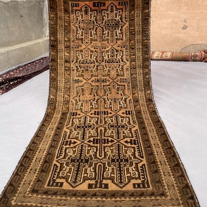 May include: A rectangular hand-knotted rug featuring a repeating geometric pattern in shades of dark brown, tan, and cream. The rug has a wide border with a repeating stepped motif and short, light-coloured fringe along the top and bottom edges.