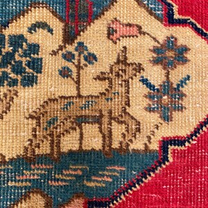 May include: A woven textile featuring a central cream-colored medallion with a deer, stylized trees, and floral motifs in shades of blue, brown, and pink. The medallion is set against a vibrant red background with dark blue decorative borders.