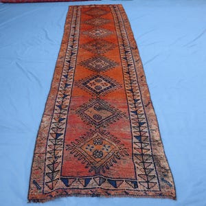 Op de afbeelding: Een oranje en rode loper met een geometrisch ruitpatroon. Het tapijt heeft een centraal ontwerp van herhalende ruiten in verschillende maten, met een rand van driehoeken en andere vormen. Geplaatst op een blauw oppervlak.