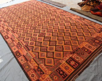 May include: A large, rectangular rug with a geometric diamond pattern in shades of brown, orange, and black. The rug has a detailed border with repeating geometric shapes. The rug is made of woven material and is suitable for home decor.