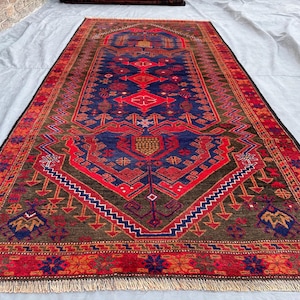 May include: A rectangular, hand-woven rug with a central blue field and intricate red and brown geometric patterns. The rug features a detailed border and fringe, showcasing a traditional design. The color palette includes shades of red, blue, brown, and touches of gold.