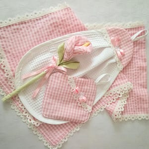 Puede incluir: Un juego de toallas de cocina de vichy rosa y blanco con ribete de encaje. Las toallas tienen una flor blanca de ganchillo y una cinta rosa.