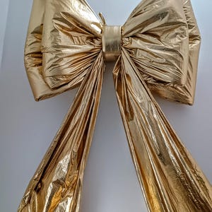 Può includere: Un grande fiocco dorato con lunghe code fluenti. Il fiocco è realizzato in un materiale metallico lucido, probabilmente per la decorazione. Il fiocco è centrato e sembra essere appeso contro un muro bianco. La finitura metallica del fiocco cattura la luce.