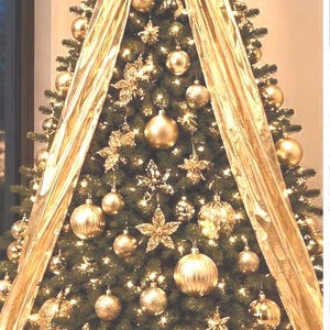 May include: A decorated Christmas tree with gold ornaments, a large bow at the top, and gold ribbon cascading down. The tree is adorned with gold baubles, star-shaped ornaments, and lights, creating a festive holiday display.
