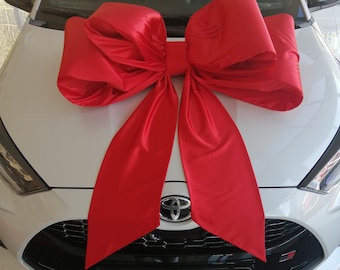 May include: A white Toyota Yaris car with a large red satin bow on the hood. The car's front features a black grille, the Toyota emblem, and the name "Yaris" on the license plate.