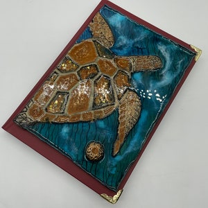 Puede incluir: Un diario encuadernado en cuero rojo con un diseño de esquina de metal dorado. La portada presenta una imagen cubierta de resina de una tortuga marina nadando en agua azul con acentos de purpurina dorada.