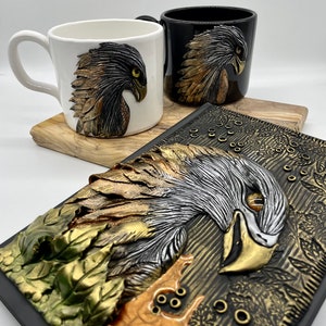 May include: Two ceramic mugs with a 3D eagle design. One mug is white with a gold and black eagle, the other is black with a gold and silver eagle. A black journal with a gold and silver eagle design is in the background.