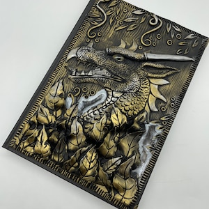 Puede incluir: Un diario de cuero negro con un diseño de dragón en 3D. El dragón está hecho de pintura dorada y plateada y tiene una apariencia texturizada y escamosa. El diario está decorado con hojas y vides doradas y plateadas.