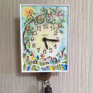 May include: A white square clock with a painted scene of a tree with red apples, houses, and a cat. The clock face is yellow with black hands and black Roman numerals. The clock is hanging from a chain with a bell.