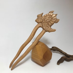 May include: A wooden hair stick with a carved turtle design. The stick is brown and has a natural finish. It is resting on a wooden block.