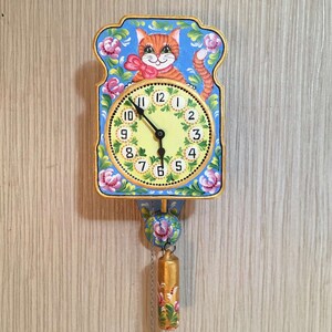 Pode incluir: Um relógio pintado de azul e dourado com um rosto de gato e detalhes florais. O mostrador do relógio é amarelo com números e ponteiros pretos. O relógio tem um pêndulo com um sino dourado.
