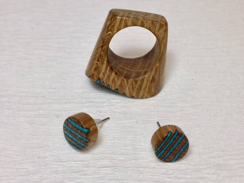 Puede incluir: Un anillo de madera y un par de pendientes con incrustaciones de turquesa. El anillo es una banda gruesa con forma cuadrada y un gran agujero en el centro. Los pendientes son redondos y tienen un patr&oacute;n de rayas turquesa.