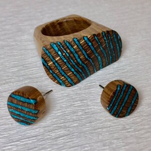 Puede incluir: Un conjunto de anillo y pendientes de madera con un dise&ntilde;o de incrustaciones de turquesa. El anillo es de forma ovalada y los pendientes son redondos. La madera es de color marr&oacute;n oscuro y la incrustaci&oacute;n de turquesa es de color azul brillante.