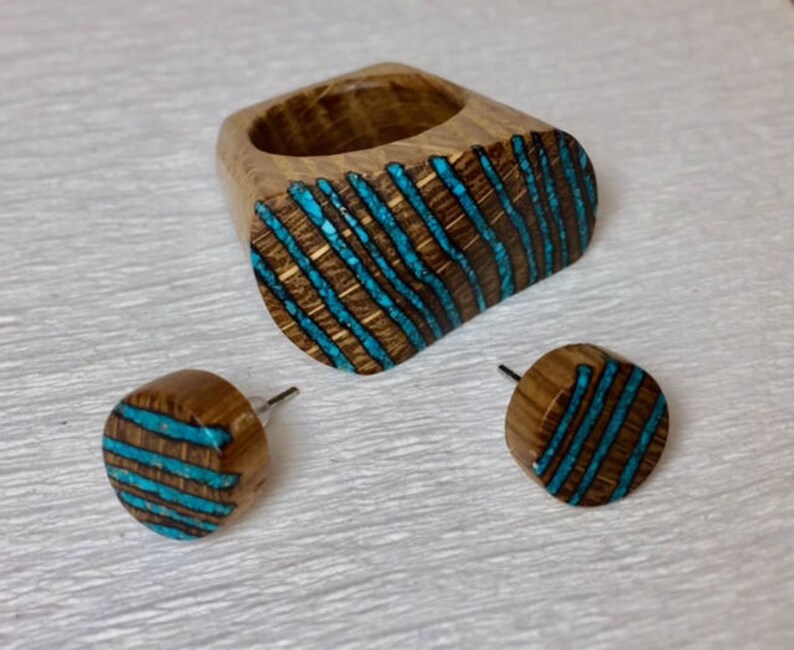 Puede incluir: Un conjunto de anillo y pendientes de madera con un dise&ntilde;o de incrustaciones de turquesa. El anillo es de forma ovalada y los pendientes son redondos. La madera es de color marr&oacute;n oscuro y la incrustaci&oacute;n de turquesa es de color azul brillante.