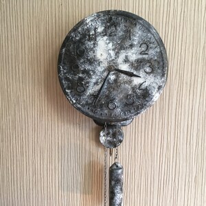 May include: A black and white pendulum clock with a distressed finish. The clock face has Roman numerals and the pendulum is hanging from the bottom of the clock.