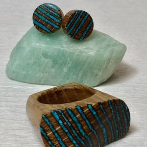 Puede incluir: Un conjunto de pendientes de madera y un anillo con incrustaciones de turquesa. Los pendientes son redondos y el anillo tiene forma ovalada. Las incrustaciones de turquesa tienen un patr&oacute;n de rayas.